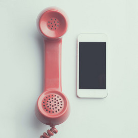 Ein altmodischer, rosafarbener Telefonhörer mit Kabel liegt neben einem weißen Smartphone. Der Hintergrund ist neutral.